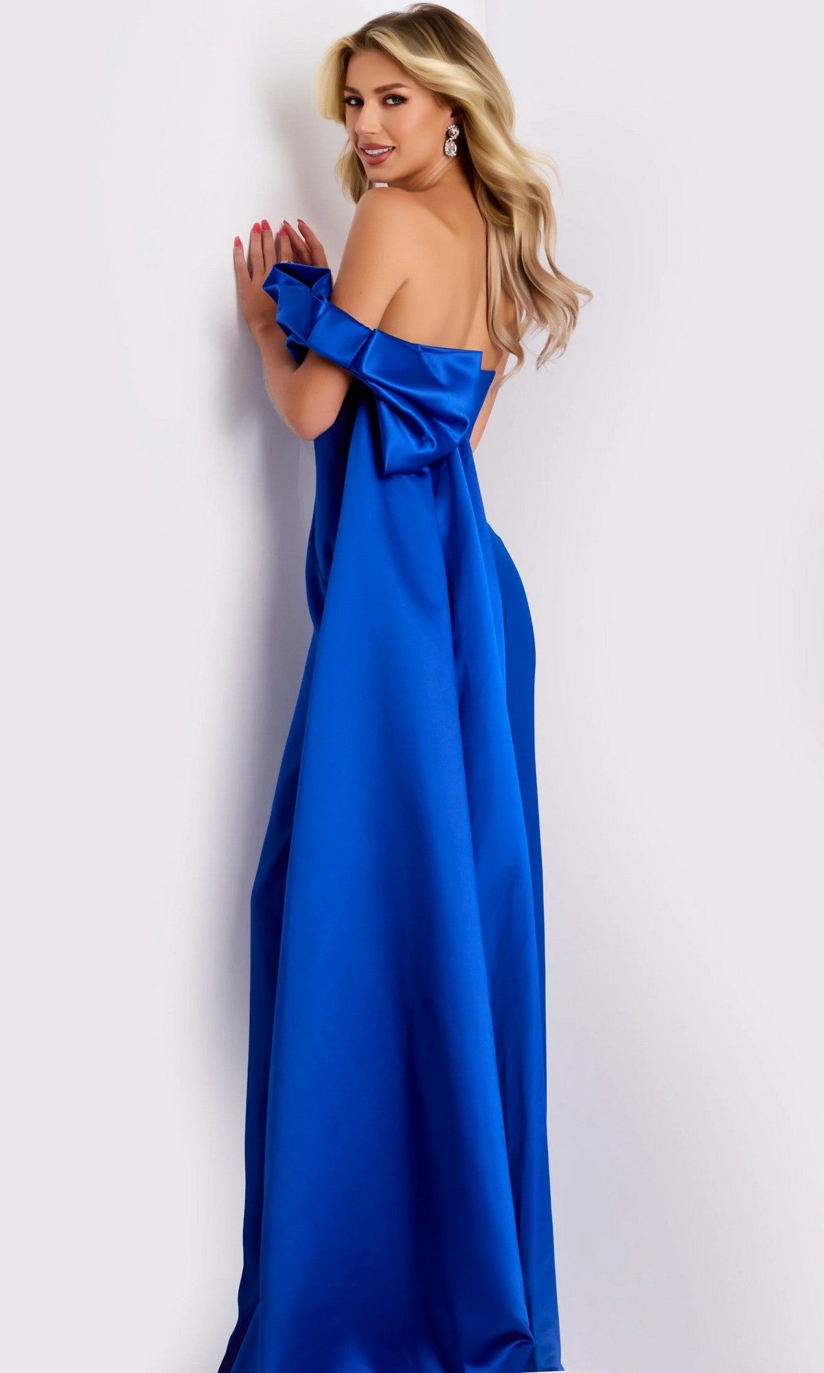 A woman in the Long Formal Dress: Jovani 38864 stands against a white background, smiling and looking over her shoulder; the strapless royal blue gown features a large bow detail.